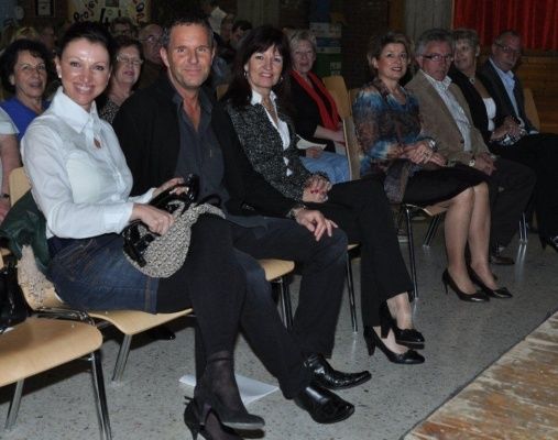 Eine Gruppe von Menschen, darunter ein Mann und eine Frau, sitzen in Stühlen in einem Raum, möglicherweise bei einer Veranstaltung. Sie schauen alle in die gleiche Richtung.