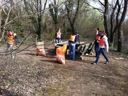Eine Gruppe von Freiwilligen in orangenen Westen räumt einen Parkbereich mit einer Holzbank und Müllsäcken auf.