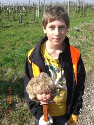 Ein junger Junge in einem gelben Shirt und oranger Weste hält eine Puppenkopf mit einem Holzstab in einem Grasfeld.