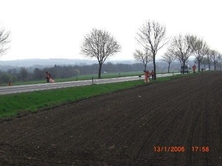 Eine ländliche Landschaft mit einem gepflügten Feld, einer Straße und mehreren Personen in roter Kleidung am Straßenrand. Bäume säumen die Straße, und der Himmel ist bedeckt.