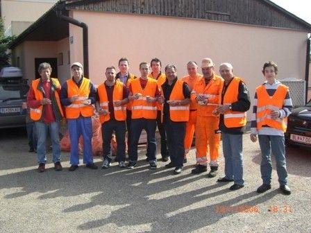 Eine Gruppe von Männern in orange Sicherheitswesten posiert für ein Foto vor einem Gebäude. Einige halten Bänder und einer trägt einen Hut. Autos sind in der Nähe geparkt.