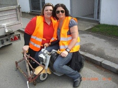 Zwei Frauen in orange Sicherheitswesten sitzen auf einem gelben Roller, eine hält das Lenkrad, die andere hat eine Tasche auf dem Boden, an einem sonnigen Tag.