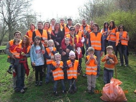 Eine Gruppe von Menschen, darunter Erwachsene und Kinder, steht auf einer Rasenfläche, trägt orange Westen und Handschuhe, und einige halten Müllsäcke. Sie scheinen an einer Gemeinschaftsaktion zur Müllbeseitigung teilzunehmen.