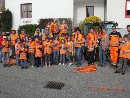 Eine Gruppe von Menschen in orangefarbenen Westen, einschließlich Kindern, steht vor einem Gebäude mit einem Traktor und einem Schild, das 'Reinwalder' besagt. Sie halten Reinigungswerkzeuge in den Händen und nehmen an einer gemeinschaftlichen Aufräumaktion teil.