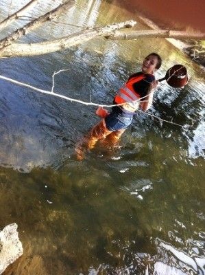 Eine Person in einem Sicherheitsweste und Stiefeln watet durch einen Fluss und hält ein Netz und einen Korb.