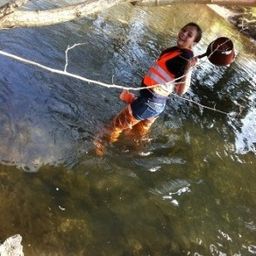Eine Person in einem Sicherheitsweste und Stiefeln watet durch einen Fluss und hält ein Netz und einen Korb.