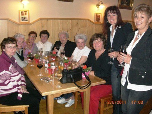 Eine Gruppe von Frauen sitzt um einen Tisch herum, lächelt und hält Weingläser in den Händen. Sie sind in verschiedenen Outfits gekleidet, und einige tragen Brillen. Auf dem Tisch stehen Blumen und eine Handtasche. Das Foto wurde am 5. März 2011 um 17:03 aufgenommen.