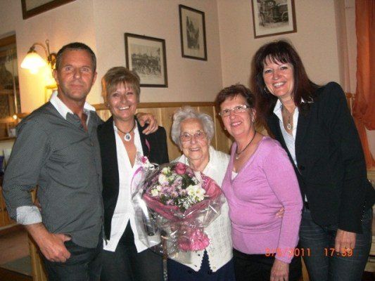 Fünf Personen posieren für ein Foto im Innenraum. Eine Frau in der Mitte hält einen Blumenstrauß. Die Frau rechts trägt eine Brille. Hinter ihnen hängen drei gerahmte Bilder an der Wand.