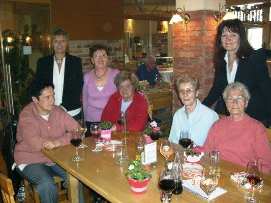 Eine Gruppe älterer Frauen und Männer sitzt in einem Restaurant um einen Tisch herum, lächelt und posiert für ein Foto. Auf dem Tisch stehen Weingläser, Topfpflanzen und eine Speisekarte. Im Hintergrund ist eine Ziegelwand und ein Glasfenster zu sehen.