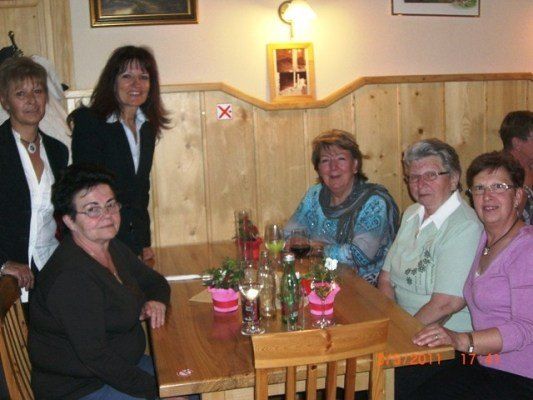 Eine Gruppe von Frauen sitzt an einem Tisch in einem Restaurant, lächelt und posiert für ein Foto. Der Tisch hat Blumen, Gläser und Flaschen. Die Wand hinter ihnen hat Bilderrahmen und ein rotes Kreuz-Schild. Das Bild hat einen Zeitstempel von 17:44.