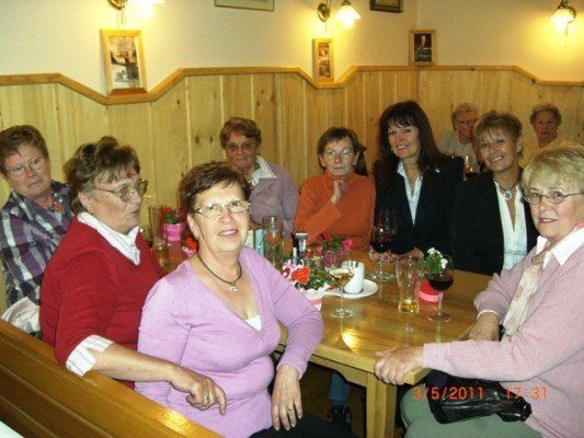 Eine Gruppe von Frauen sitzt um einen Holztisch in einem Restaurant, lächelt und posiert für ein Foto. Auf dem Tisch stehen Blumen, Weingläser und Teller. Die Wände sind aus Holz und es hängen Bilderrahmen daran. Das Datum und die Uhrzeit sind in der unteren rechten Ecke des Bildes angezeigt.