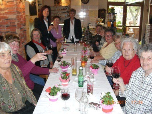 Eine Gruppe älterer Frauen sitzt um einen langen Tisch herum, hält Weingläser in der Hand und lächelt für ein Foto. Der Tisch ist mit kleinen Topfpflanzen und Weingläsern geschmückt. Eine Frau in einem schwarzen Anzug steht hinter ihnen.