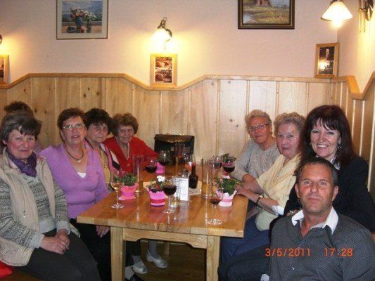 Eine Gruppe von Menschen, darunter Frauen und Männer, sitzt um einen Holztisch in einem Restaurant. Sie lächeln und posieren für ein Foto. Der Tisch hat Weingläser, Blumen und eine Flasche. Die Wand hat Holzpaneele und Bilderrahmen. Das Datum und die Uhrzeit sind 3/5/2011 17:28.