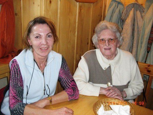 Zwei Frauen sitzen an einem Tisch, eine lächelt, die andere trägt eine Brille, mit einer hölzernen Wand und hängenden Kleidern im Hintergrund.