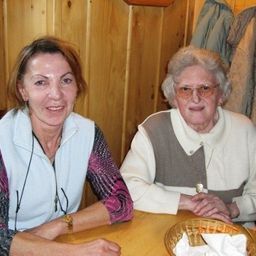 Zwei Frauen sitzen an einem Tisch, eine lächelt, die andere trägt eine Brille, mit einer hölzernen Wand und hängenden Kleidern im Hintergrund.