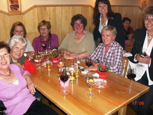 Eine Gruppe von Frauen sitzt um einen Tisch mit Weinglas und Blumen und lächelt für ein Foto. Der Tisch hat einen Teller, eine Kerze und eine Topfpflanze. Der Hintergrund ist eine Holzwand.