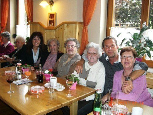 Eine Gruppe älterer Menschen, darunter Frauen und Männer, versammelt sich in einem Restaurant um einen Tisch, lächelt und posiert für ein Foto. Der Tisch ist mit Weingläsern, Tellern und verschiedenen Getränken gedeckt. Im Hintergrund ist eine Pflanze zu sehen.
