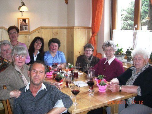 Eine Gruppe älterer Menschen, sowohl Männer als auch Frauen, sitzen um einen Tisch in einem Restaurant, lächeln und posieren für ein Foto. Der Tisch ist mit Weingläsern, Topfpflanzen und anderen Gegenständen gefüllt. Der Raum hat Holzwände und ein Fenster mit Vorhängen.