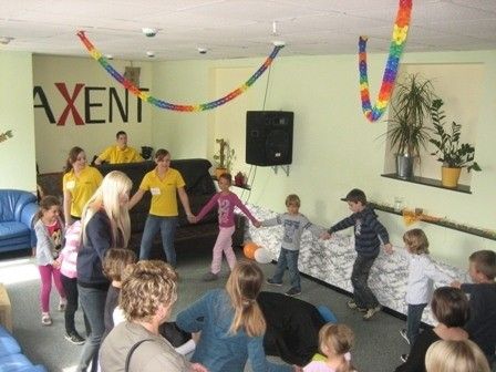 Eine Gruppe von Kindern und Erwachsenen tanzt in einem Raum mit einer Couch, einem Lautsprecher und Topfpflanzen an der Wand. Das Wort 'AXENT' ist an der Wand geschrieben.