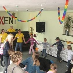 Eine Gruppe von Kindern und Erwachsenen tanzt in einem Raum mit einer Couch, einem Lautsprecher und Topfpflanzen an der Wand. Das Wort 'AXENT' ist an der Wand geschrieben.