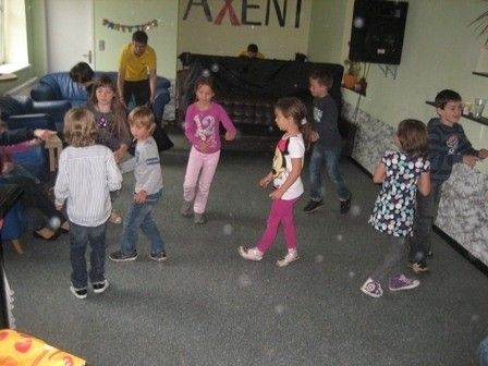 Eine Gruppe von Kindern tanzt in einem Raum mit Teppichboden. Eine Frau in einem gelben Shirt steht in der Mitte. Ein Sofa und ein Fernseher sind im Hintergrund.