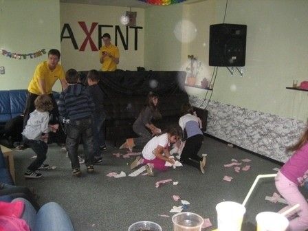Eine Gruppe von Kindern und Erwachsenen spielt in einem Raum mit einem schwarzen Sofa, verstreuten Papieren und Bechern auf dem Boden. Ein Mann in einem gelben Shirt steht in der Nähe des Sofas. Ein Lautsprecher ist an der Wand montiert.