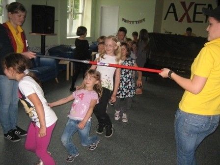Eine Gruppe von Kindern und Erwachsenen spielt ein Spiel mit einem langen roten Stock in einem Raum mit blauen Stühlen und einer Wand mit dem Wort Axem.