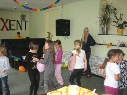 Eine Gruppe von Kindern spielt mit Luftballons in einem Raum mit einer DJ-Bühne, einer stehenden Frau und Topfpflanzen an der Wand.