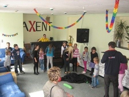 Eine Gruppe von Kindern und Erwachsenen versammelt sich in einem Raum, einige stehen und andere sitzen auf einer Couch. Ein Mann steht vor ihnen und an der Wand befindet sich ein Lautsprecher. Der Raum ist mit bunten Girlanden und Pflanzen dekoriert.