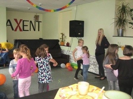 Eine Gruppe junger Mädchen tanzt in einem Raum mit einer gelben Tischdecke, Luftballons und einem Lautsprecher. Eine Frau steht in der Nähe. An der Wand steht das Wort 'AXENT'.