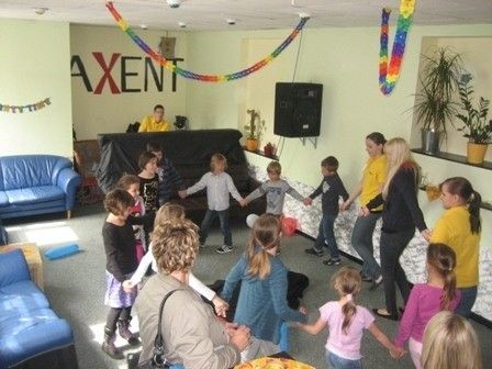 Eine Gruppe von Kindern und Erwachsenen nimmt an einer Tanzaktivität in einem Raum mit blauen Sofas, einem schwarzen Sofa und Topfpflanzen teil. Der Raum hat ein Banner mit dem Wort 'AXENT' an der Wand.