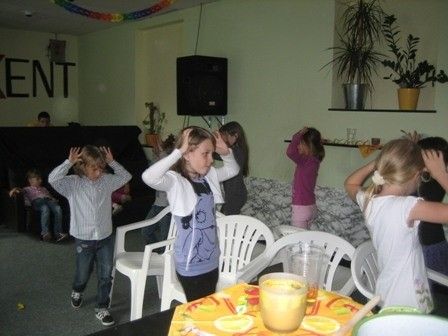 Eine Gruppe von Kindern ist drinnen, einige stehen und andere sitzen, wobei ein Mädchen einen Becher mit Orangensaft hält. Ein Lautsprecher ist an der Wand montiert und Topfpflanzen stehen auf einem Regal.
