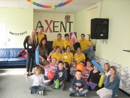 Eine Gruppe von Erwachsenen und Kindern posiert für ein Foto in einem Raum mit einer blauen Couch, Luftballons und einem Lautsprecher. Die Erwachsenen tragen gelbe Hemden mit dem Wort Axent darauf.