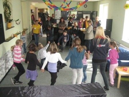 Eine Gruppe von Kindern und Erwachsenen versammelt sich in einem Raum, einige tanzen, während andere zuschauen. Der Raum ist mit bunten Bändern und Luftballons dekoriert. Es gibt Topfpflanzen, einen Tisch mit Essen und eine Couch im Hintergrund.