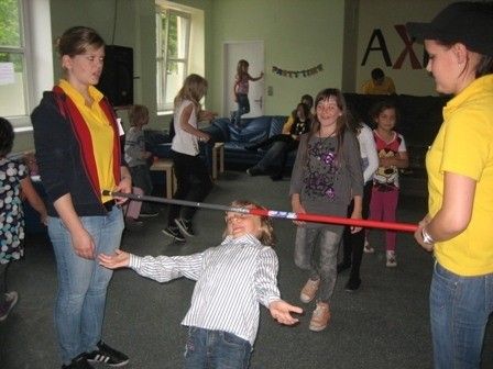 Eine Gruppe von Kindern und Erwachsenen befindet sich in einem Raum. Ein Kind liegt mit einem Stock in der Hand auf dem Boden. Zwei Erwachsene halten einen langen roten Stock.