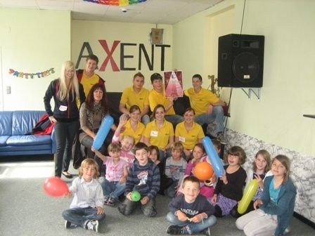 Eine Gruppe von Menschen, einschließlich Erwachsener und Kindern, posiert für ein Foto in einem Raum mit einem blauen Sofa, Luftballons und einem Lautsprecher. An der Wand steht das Wort AXENT.