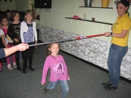 Eine Frau hält einen langen rot-weißen Stab, während ein junges Mädchen in einem rosa Shirt versucht, ihn ihr zu entreißen. Andere Kinder beobachten im Hintergrund.