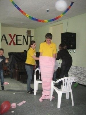 Eine Gruppe von Menschen versammelt sich in einem Raum, zwei von ihnen halten ein großes rosa Objekt, möglicherweise ein Ballon, während andere stehen und zusehen. Der Raum ist mit bunten Dekorationen und Möbeln ausgestattet.