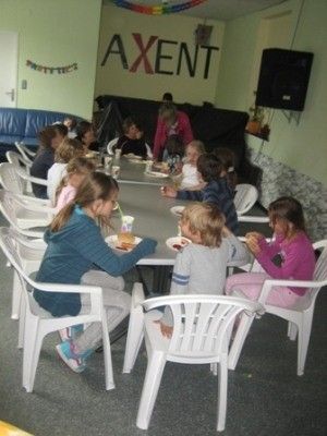 Eine Gruppe von Kindern sitzt um einen Tisch in einem Raum und isst. Eine Frau steht hinter ihnen. Der Raum hat ein Banner mit dem Wort 'AXENT' und einen montierten Fernseher.