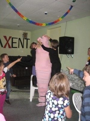 Eine Gruppe von Kindern spielt mit einer großen rosa Figur in einem Raum mit einem weißen Stuhl und einem Lautsprecher an der Wand.