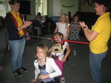 Eine Gruppe von Kindern und Erwachsenen ist drinnen, einige halten einen langen roten Stab. Eine Erwachsene klatscht, und die Kinder tragen verschiedene Kleidungsstücke.