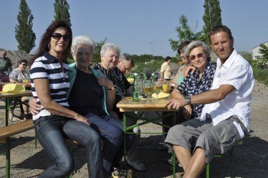 Eine Gruppe von Menschen, darunter ältere Frauen und Männer, sitzen an einem Tisch in einer Outdoor-Umgebung, möglicherweise bei einem Picknick oder Gartenfest. Sie lächeln und scheinen sich zu amüsieren.
