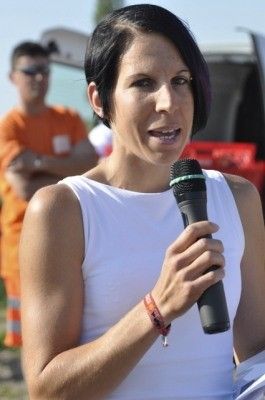 Eine Frau in einem weißen ärmellosen Oberteil hält ein Mikrofon und scheint bei einer Veranstaltung im Freien zu sprechen, wobei der Hintergrund einen verschwommenen Menschen in einem orangefarbenen Shirt zeigt.