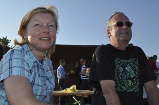 Eine Frau in einem blauen karierten Shirt und ein Mann in einem schwarzen T-Shirt mit Baumdesign lächeln und sitzen zusammen bei einer Veranstaltung im Freien. Hinter ihnen ist eine Holzstruktur und mehrere Personen zu sehen.