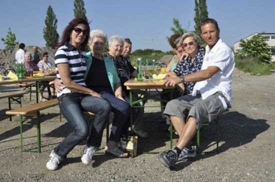 Eine Gruppe von Menschen, darunter ältere Frauen und ein Mann, sitzt an einem Picknicktisch im Freien. Sie lächeln und scheinen eine Mahlzeit zu genießen. Der Tisch ist mit Flaschen, Gläsern und Essen gedeckt. Hinter ihnen sitzen weitere Menschen an anderen Tischen. Bäume und ein Gebäude sind im Hintergrund zu sehen.