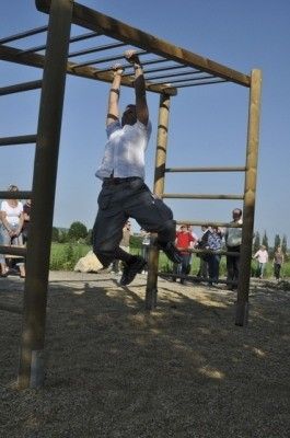 Ein Mann führt Klimmzüge an einem hölzernen Klimmzuggerät in einer Außenumgebung durch, während mehrere Personen im Hintergrund zusehen.
