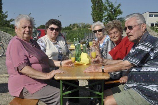 Eine Gruppe älterer Menschen sitzt um einen Tisch herum, lächelt und hält Getränke. Sie sind draußen in einem Park.