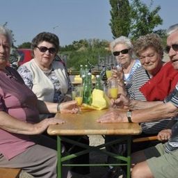Eine Gruppe älterer Menschen sitzt um einen Tisch herum, lächelt und hält Getränke. Sie sind draußen in einem Park.