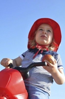 Ein junges Kind in einem roten Hut und gestreiftem Shirt fährt ein rotes Spielzeugmotorrad und hält einen Apfel in der Hand.