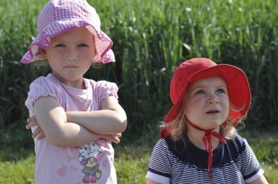 Zwei kleine Mädchen stehen in einem Feld, eines mit verschränkten Armen und das andere mit einem roten Hut. Sie sehen ernst aus.